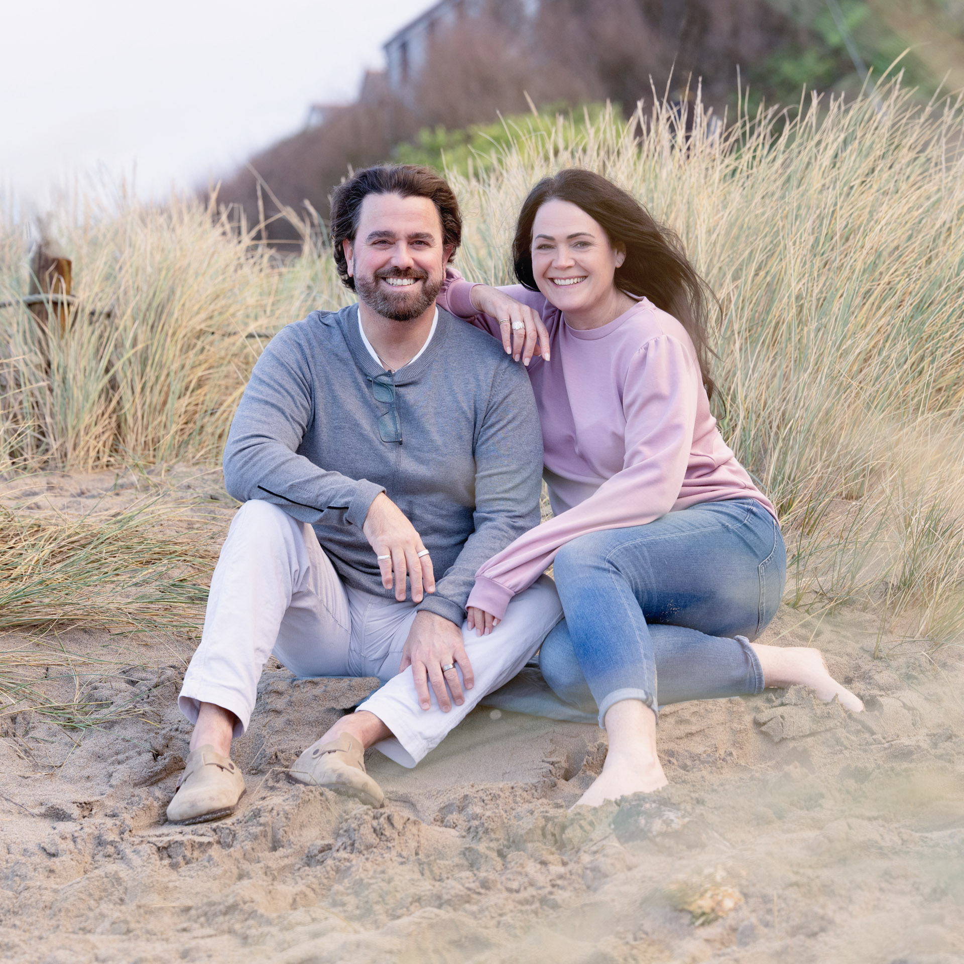 Smiling couple sitting together on sandy dunes with tall grass, relaxed and close, in a calm coastal setting.