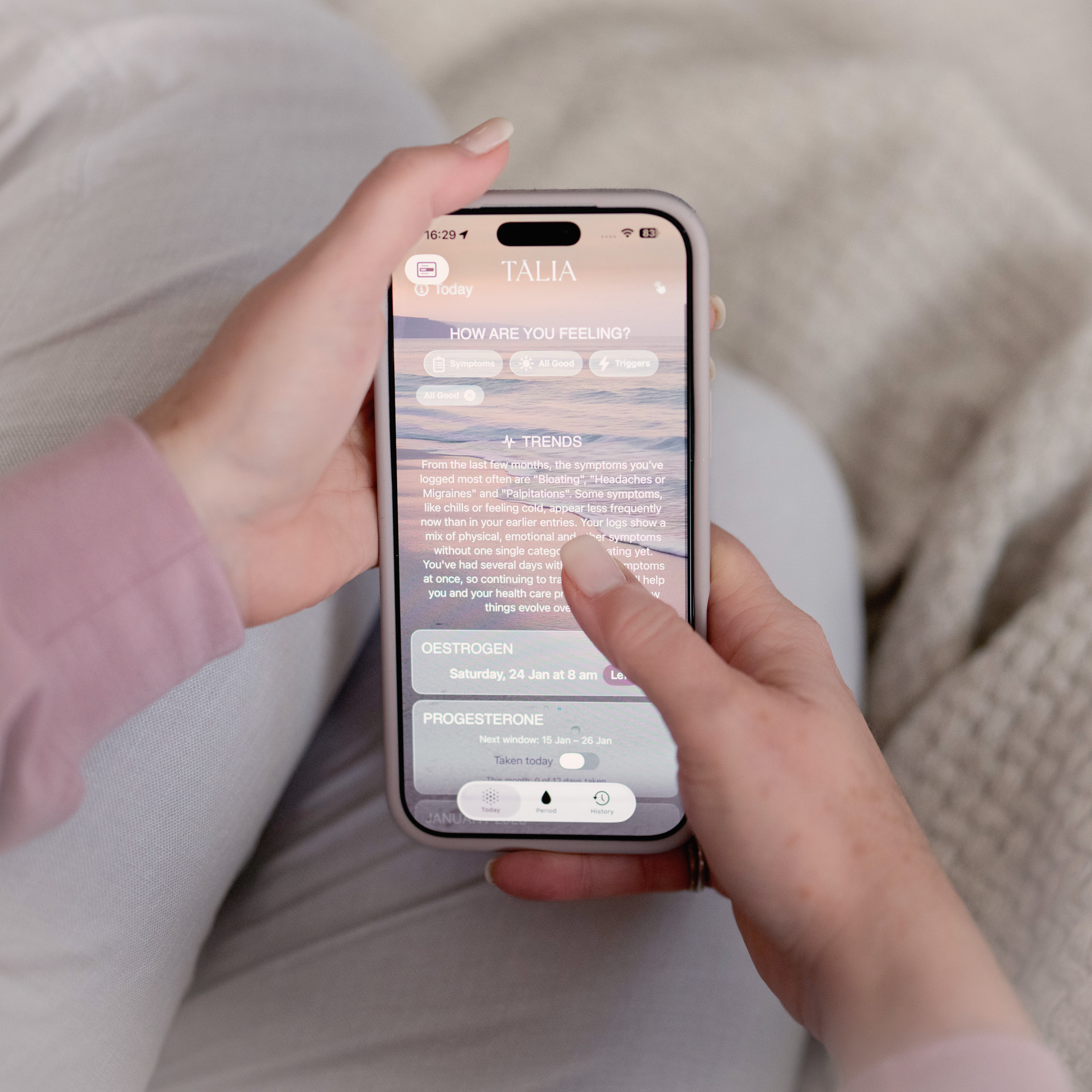 Close-up of a person holding an iPhone displaying the TĀLIA app, showing symptom trends and HRT tracking information, with a soft, neutral background.