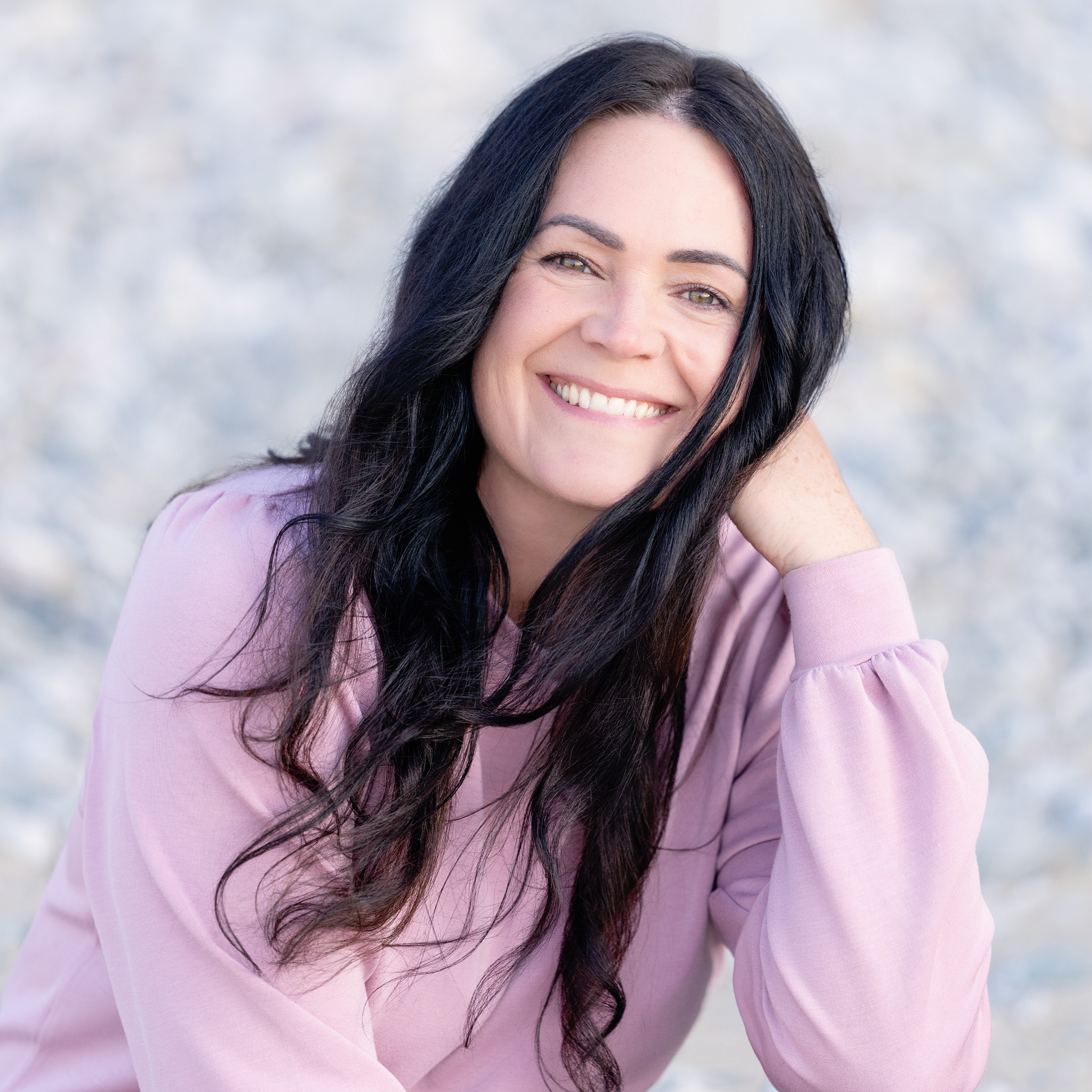 Close-up portrait of a smiling woman with long dark hair wearing a pink sweater, blurred light background.